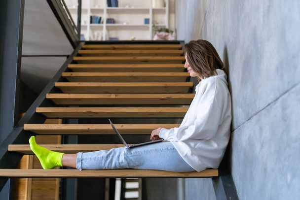 Μια νεαρή γυναίκα στο σπίτι με λευκή κουκούλα και τζιν κάθεται στις σκάλες με ένα λάπτοπ στα γόνατά της, δουλεύει εξ αποστάσεως, σπουδάζει. - Φωτογραφία, εικόνα