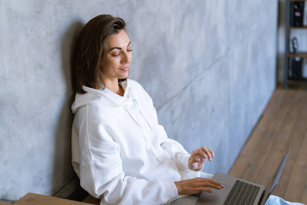 Una joven mujer en casa con una sudadera blanca se sienta en las escaleras pensativamente soñadora mira, sostiene una computadora portátil en sus rodillas - Foto, Imagen