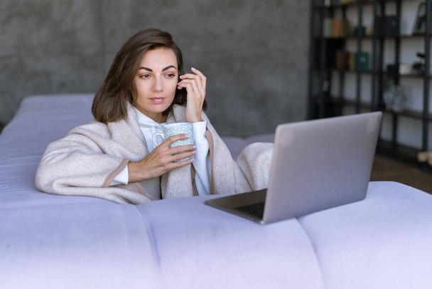 Young woman at home on the sofa in winter under a cozy blanket with a laptop, watching a movie, TV series, reading a book on a laptop - Foto, afbeelding
