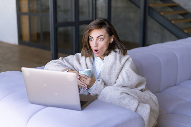 Νεαρή γυναίκα στο σπίτι στον καναπέ το χειμώνα κάτω από μια ζεστή κουβέρτα με ένα φορητό υπολογιστή, σοκαρισμένη κοιτάζει την οθόνη - Φωτογραφία, εικόνα
