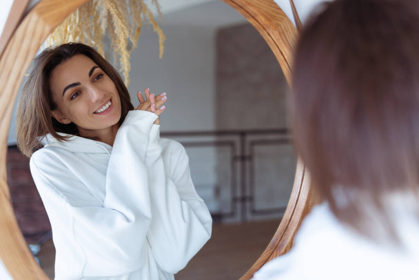 Молода жінка вдома в спальні в теплому білому светрі позує перед дзеркалом
 - Фото, зображення