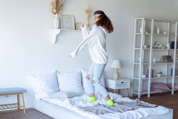 Mujer joven en casa en el dormitorio en una sudadera con capucha blanca cálida, divertirse, saltar en la cama, bailar - Foto, Imagen