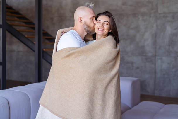 Νεαρό ζευγάρι άντρας και γυναίκα στο σπίτι αγκαλιάζονται κάτω από μια ζεστή κουβέρτα, αγκαλιάζονται, δείχνουν τρυφερά συναισθήματα - Φωτογραφία, εικόνα