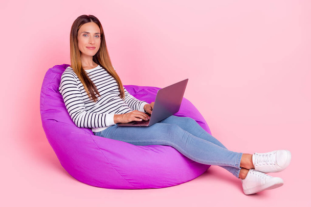 Photo of pretty shiny young woman dressed striped pullover sitting bean bag working modern device smiling isolated pink color background - Foto, Bild