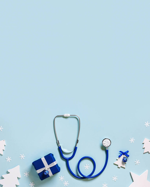 Christmas medical background with copy space, blue stethoscope, gift box, christmas ball, snowflakes, New Year wooden toy fir tree. Winter medicine concept for business. Vertical image - Fotoğraf, Görsel