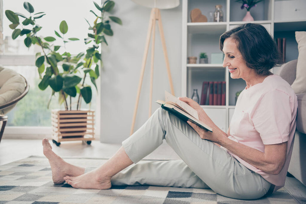 Full length body size side profile photo senior woman sitting on floor in casual clothes reading book spending free time - 写真・画像