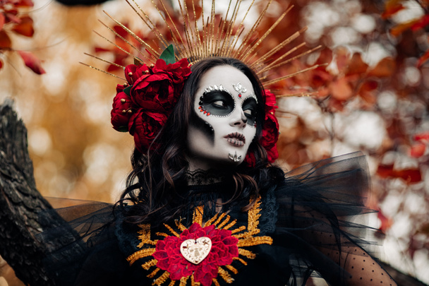 Portrait of young woman with sugar skull makeup and red roses dressed in black costume of death as Santa Muerte against background of autumn leaves in forest. Day of the Dead or Halloween concept. - Photo, image