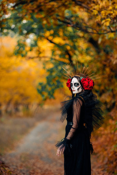 Giovane donna con trucco teschio di zucchero e rose rosse vestite in costume nero della morte come Santa Muerte è sullo sfondo della foresta autunnale. Giorno dei morti o concetto di Halloween. - Foto, immagini