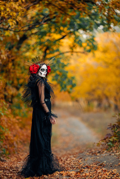 Fiatal nő a cukor koponya smink és vörös rózsák öltözött fekete jelmez a halál, mint Santa Muerte hátterében őszi erdő. Day of the Dead vagy Halloween koncepció. - Fotó, kép