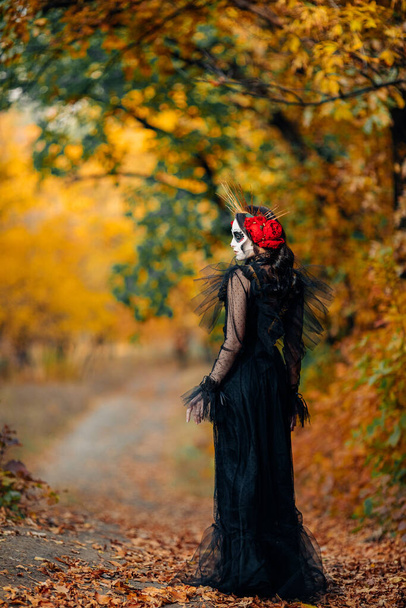 Young woman with sugar skull makeup and red roses dressed in black costume of death as Santa Muerte is against background of autumn forest. Day of the Dead or Halloween concept. - Photo, Image