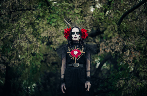 Young woman with sugar skull makeup and red roses dressed in black costume of death as Santa Muerte is against background of forest. Day of the Dead or Halloween concept. - Photo, Image