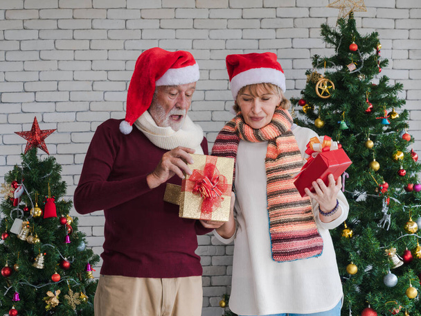 サンタ帽子の幸せな白人のシニアカップルは、装飾されたクリスマスツリーで家で一日中クリスマスプレゼントを交換することに集中しています。クリスマスと新年のお祝い活動. - 写真・画像