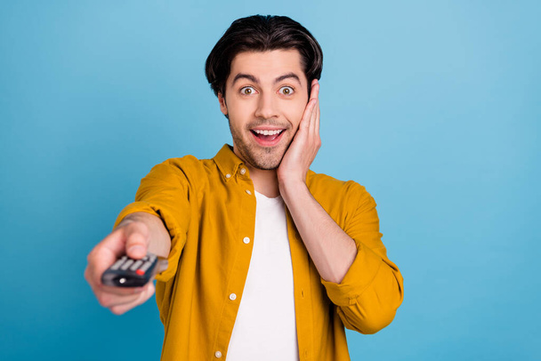 Photo of excited funny guy hold remote control watch movie palm cheek wear yellow shirt isolated blue color background - Φωτογραφία, εικόνα