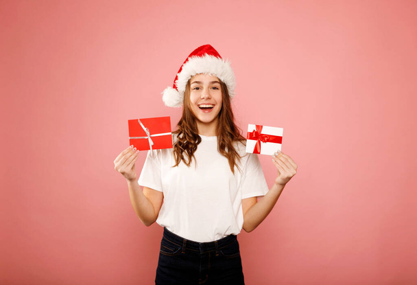 Happy caucasian girl teenager standing in white t-shirt and santa claus red hat holding two gift certificates or greeting inviting cards over pink background. Studio portrait. Christmas and New year Holiday concept. - Photo, image