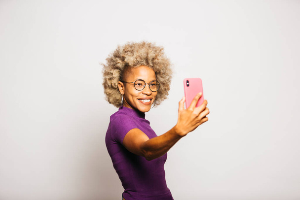 Портрет улыбающейся молодой женщины с помощью смартфона, стоя на белом фоне - Фото, изображение