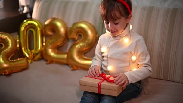 egy boldog lány karácsonyi rénszarvas fejpánttal kinyitja az ajándékdobozt. a háttérben arany számok 2022. koncepció boldog karácsonyt - Felvétel, videó