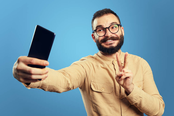Homme en tenue tendance souriant et montrant un geste de paix tout en prenant selfie - Photo, image