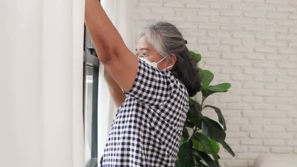 自宅に住んでいるマスクの高齢者の女性は、コロナウイルス(Covid-19)パンデミックから身を守るために窓のブラインドを開き、冷静に外を見る. - 映像、動画