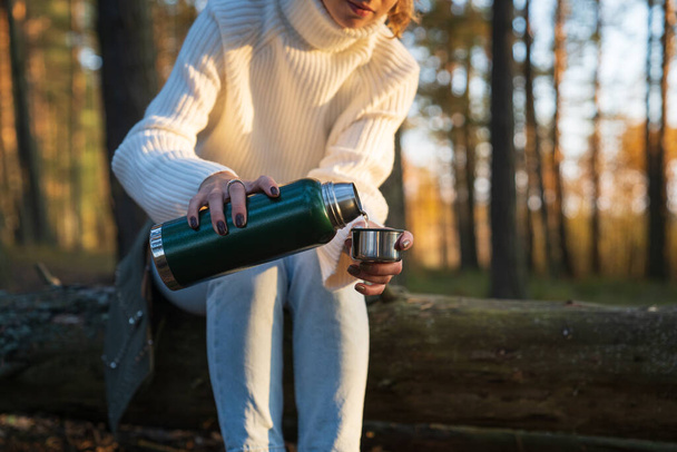 Young woman pouring hot tea from thermos in cup has rest from hike walking in autumn forest alone - Foto, Imagem