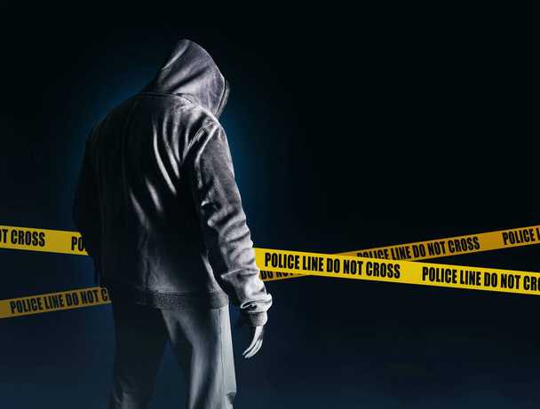 Фото страшного незнакомца-преследователя в черном капюшоне и в одежде на темно-синем фоне с полицейскими линиями. - Фото, изображение