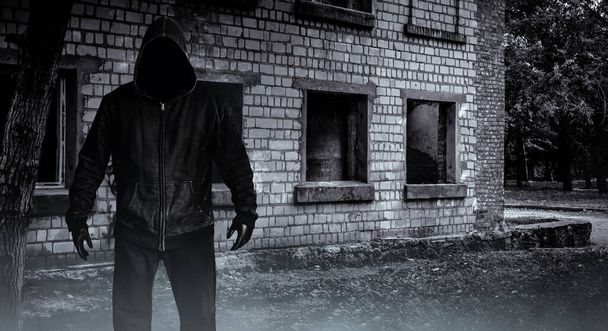 Фото страшного незнакомца-преследователя в черном капюшоне и в одежде на фоне заброшенного здания. - Фото, изображение