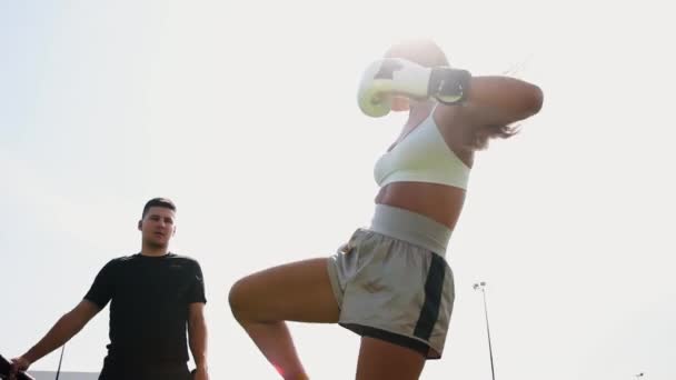 Обучение боксу на открытом воздухе - молодая женщина пинает цель в руках своих тренеров - Кадры, видео