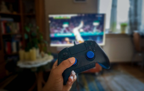 Mano humana sosteniendo un mando de juego de joystick de una consola de videojuegos contra una televisión o un televisor jugando fútbol o fútbol. Concepto de disfrute interior - Foto, Imagen
