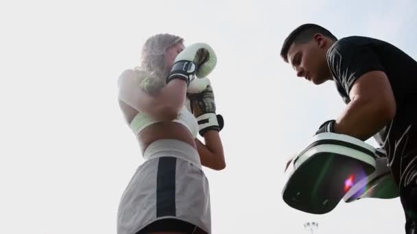 Бокс тренировки на открытом воздухе - молодая женщина с длинными волосами ногами перчатку с коленом на руках тренеров - Кадры, видео