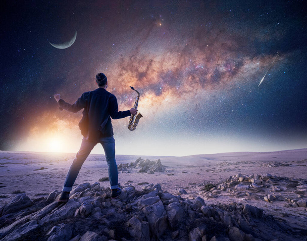 天の川の下の岩の上に腕を開いている音楽家やサックス奏者のシルエットと星空の夜、アタカマ砂漠、チリ - 写真・画像