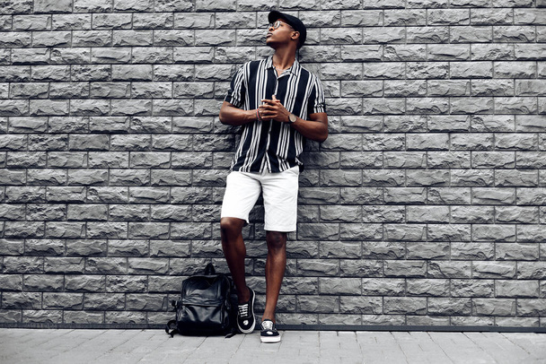 handsome african man in a cap and shirt with a backpack, against the background of a brick black wall - Φωτογραφία, εικόνα