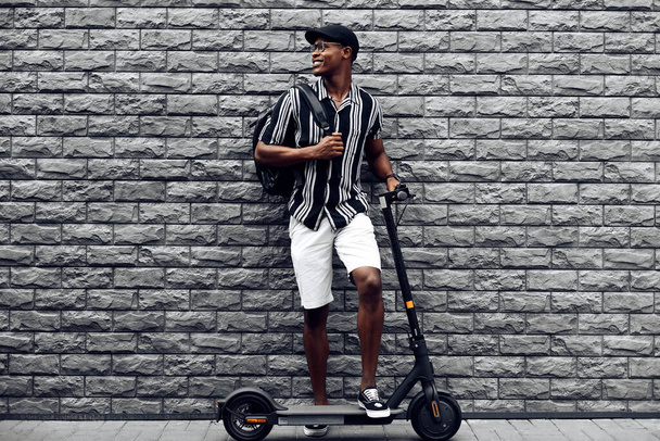 African american man with scooter on black brick wall background, guy with backpack, young student with electric scooter - Foto, immagini