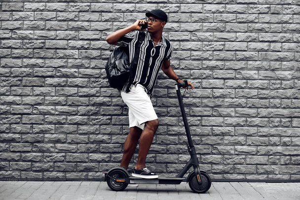 stylish african american man calling, talking on the phone, eating scooter, against black brick wall background, business meeting concept - 写真・画像
