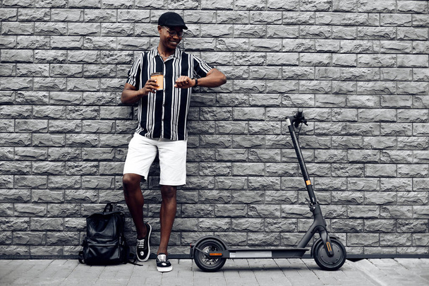 stylish african american man with paper cup of coffee or tea in hand, and electric scooter, stands against black wall background, looks at the clock on his hands, a break in working hours - Photo, Image