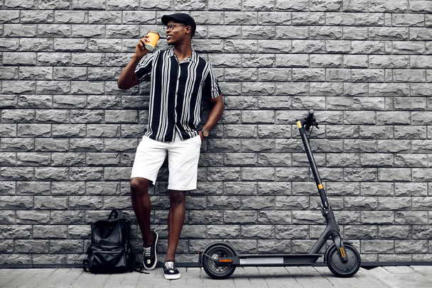 stylish african american man with paper cup of coffee or tea in hand, and electric scooter, stands against black wall background, outdoor recreation - 写真・画像