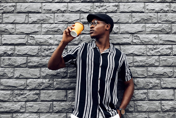 Portrait of fashionable smiling african american man in hat, man outdoors with takeaway coffee cup, over isolated gray wall background, drinks, technology and people concept, drinks from a paper cup - Fotografie, Obrázek
