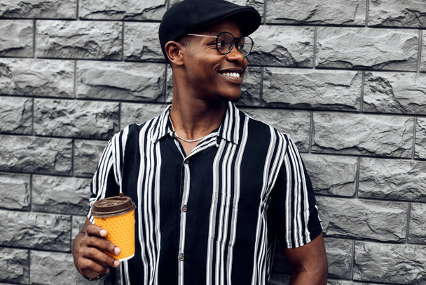 Portrait of fashionable smiling african american man in hat, man outdoors with takeaway coffee cup, over isolated gray wall background, drinks, technology and people concept - Foto, immagini
