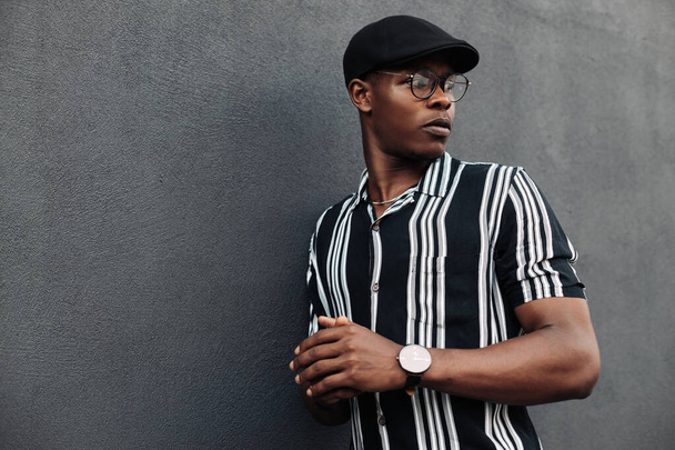 African American man in a shirt in a line, against a background of a black brick wall, stylish guy, close-up, wearing glasses, fashion posing, a black guy looks to the side, a cap - Photo, image