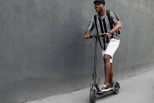 African american man with scooter on black brick wall background, guy with backpack, young student with electric scooter - Zdjęcie, obraz