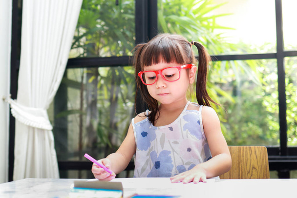 linda niña asiática con gafas rojas, de pie sobre la mesa en casa dibujos de dibujo. conceptos educativos, desarrollar el aprendizaje de los niños - Foto, Imagen