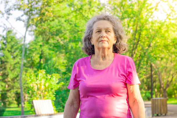 A mature woman walks in the park against the background of green tree crowns on a sunny day. An elderly woman with a serious face. Depressive mood. Psychology. Proud look. The older generation. - Photo, image