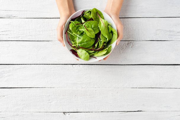 Overhead shot of woman hands holding green salad in bowl on white wooden background with copyspace - Photo, image