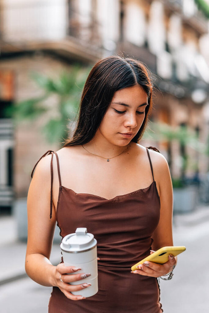 молодая китаянка держит многоразовый забрать кружку кофе и использовать ее телефон - Фото, изображение