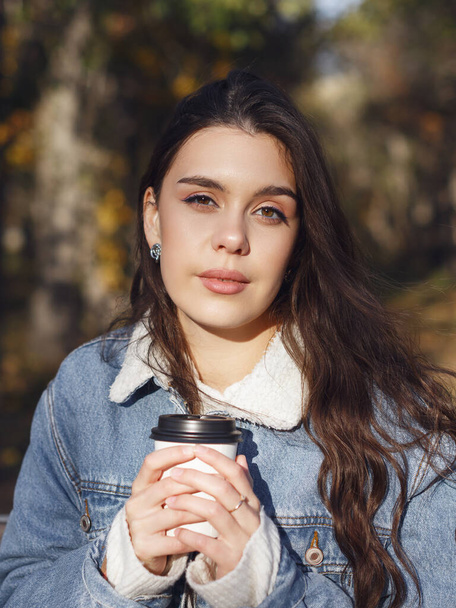 A young attractive woman is sitting on a bench in an autumn park with a cup of coffee in her hands - Foto, imagen