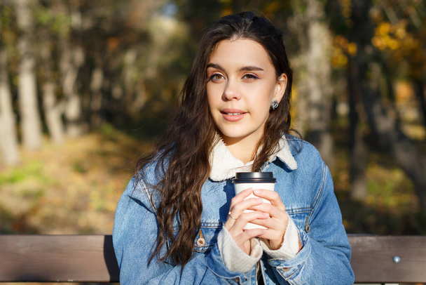 A young attractive woman is sitting on a bench in an autumn park with a cup of coffee in her hands - Photo, Image