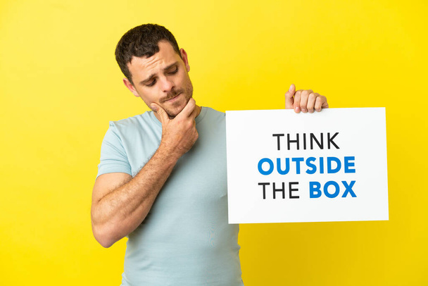 Braziliaanse man over geïsoleerde paarse achtergrond met een plakkaat met tekst Think Outside The Box en denken - Foto, afbeelding