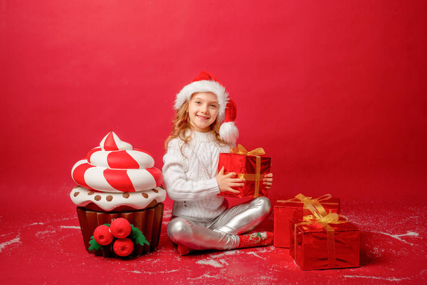 niña en un sombrero de Santa sobre un fondo rojo con una magdalena y regalos - Foto, Imagen