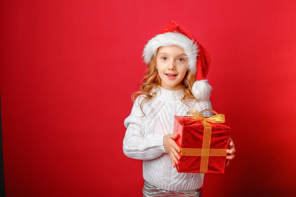 маленькая девочка в шляпе Санты на красном фоне с подарком - Фото, изображение