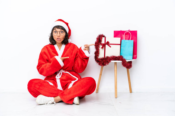 Junges Mädchen, das Weihnachten feiert, sitzt auf dem Fußboden isoliert auf weißem Bakcground überrascht und zeigt auf die Seite - Foto, Bild