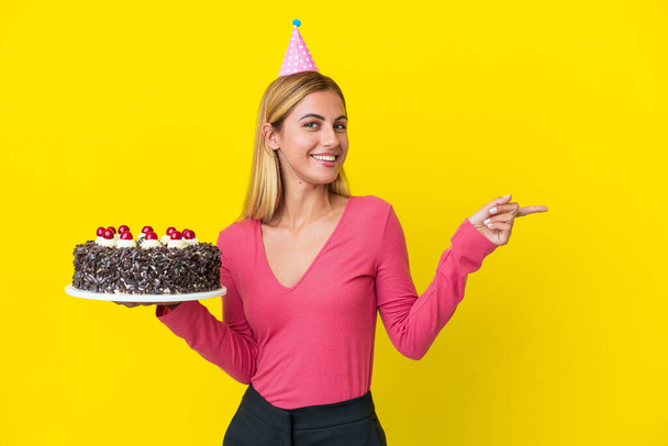 Блондинка Уругвая держит торт на день рождения изолирован на желтом фоне указывая пальцем в сторону - Фото, изображение
