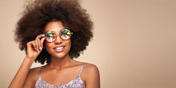 Beauty portrait of African  girl in holographic sunglasses. Beautiful black woman on beige background. Cosmetics, makeup and fashion - Photo, image